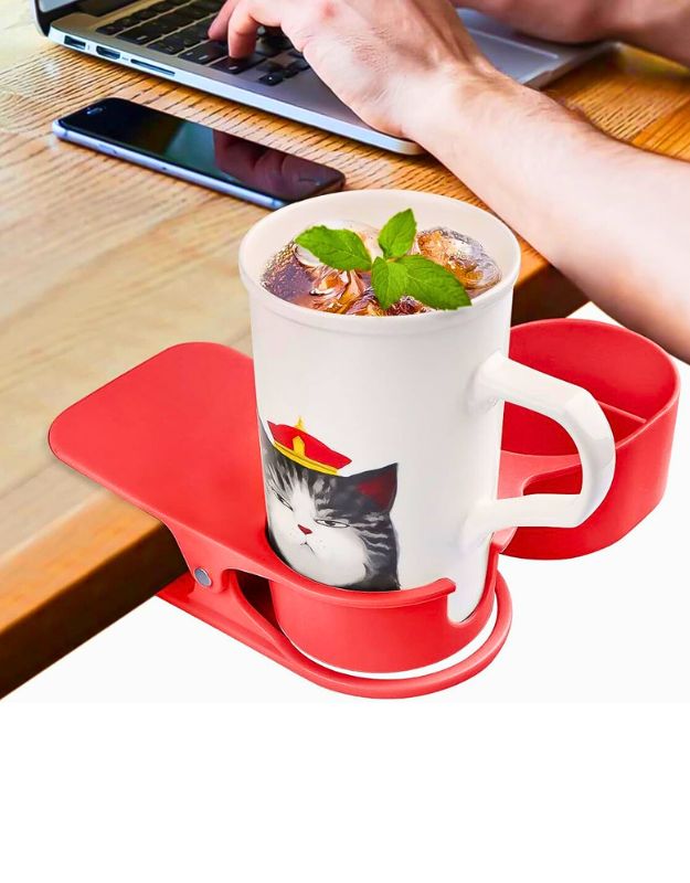 Red mug holder clip attached to a wooden desk, holding a mug with a cat design and a cold drink, near a laptop and phone.